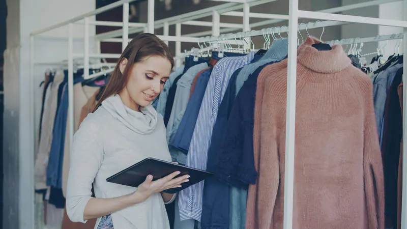 The owner of a clothing shop holds a tablet between racks of clothes.