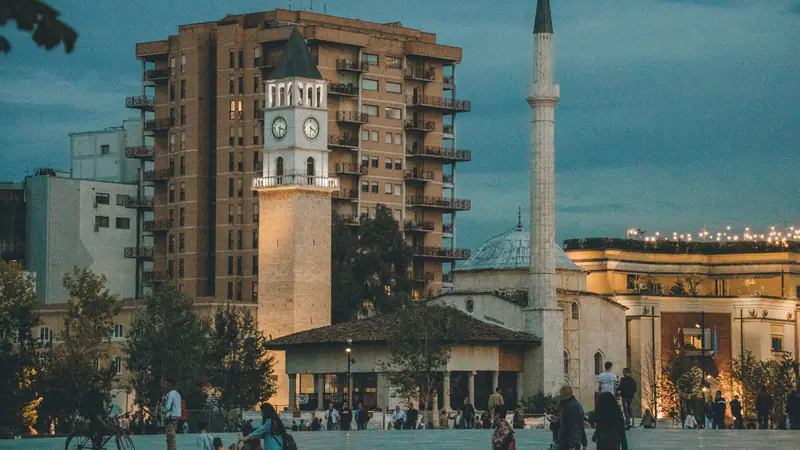 Kulla e Sahatit dhe Xhamia e Et’hem Beut në qendër të Tiranës në muzg, me njerëz që kalojnë sheshin.