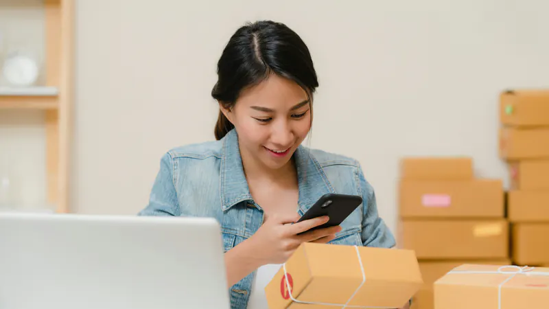 A seller photographs a parcel ready to ship with her phone, next to a laptop and more boxes.