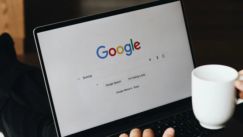 Dikush shkruan një kërkim në Google në laptop, me një filxhan kafe në dorë.